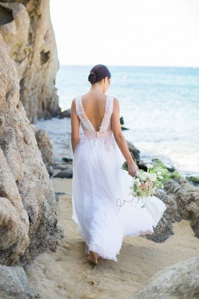 Bride with an ancient Greek-inspired wedding dress and bridal flowers walking on the coast next to grey sea rocks.