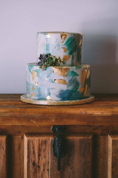 Green and gold sugar paste covered double tier wedding cake decorated with a little succulent, on a wooden surface.