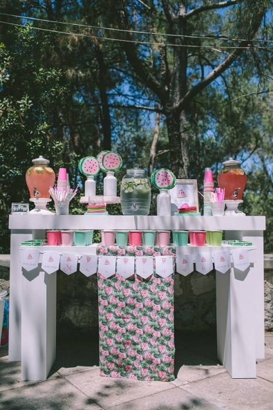 Refreshment stand with lemonade and flavored water and pastel colored paper cups, watermelon tablecloth and hand fans for a summer baptism ceremony in Greece.