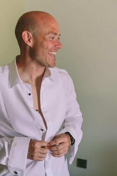 The groom putting on his white wedding shirt.