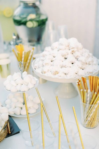 Dessert table candy bar with clear glasses with gold straws and greek almond cookies with sugar on serving trays.