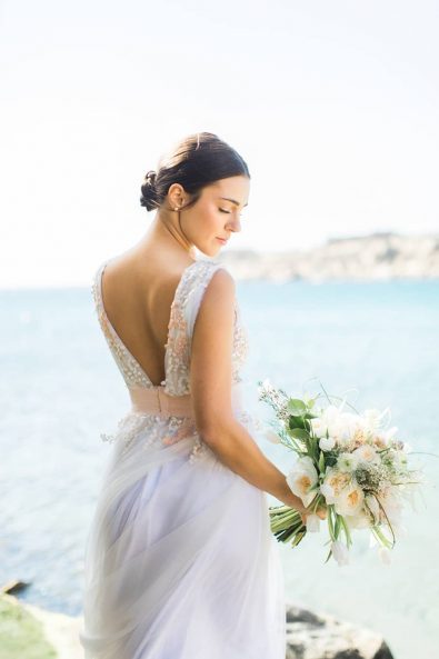 Portrait of blushing bride with an amazing backline wedding dress and low bu posing in the beachside with her flowers.