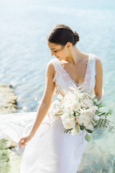 Fine art wedding photography portrait of a bride with a white and blush wedding dress and a white peonies bridal bouquet standing barefoot on the sand.