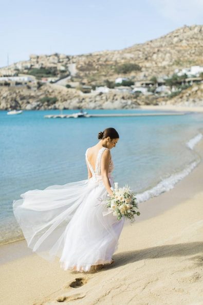 Bride walking barefoot on the mykonian beach while the breeze blows her wedding dress.