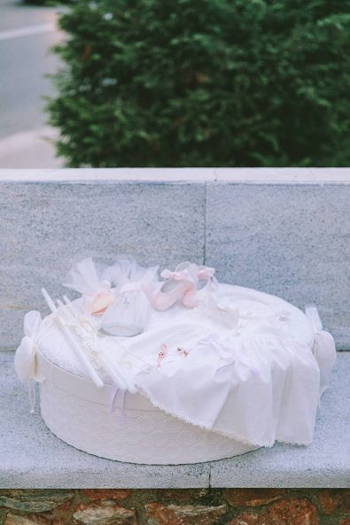 Baby's baptism ideas christening clothes on a white fabric box, lace baptism dress, pale pink ballet slippers, white baptistery candles, and oil bottle.
