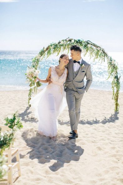 Beach wedding inspiration shoot with elegant bride and stylish groom walking on the sand in front of the wedding arch.