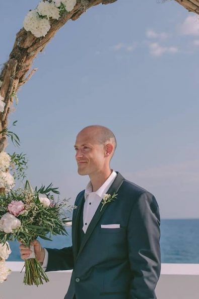 Handsome groom in a black tuxedo waiting for his bride under the wedding arch for a destination wedding in kalamata.
