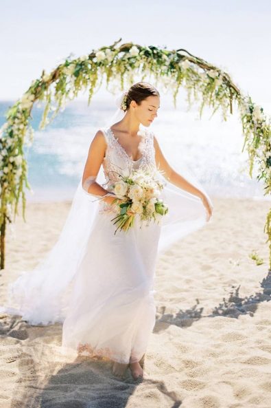 Bride standing barefoot on the beach in front of the wedding arch while the breeze caresses her tulle veil.