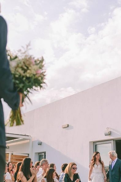 Groom holding the bridal bouquet waiting for his bride to arrive with her father at the wedding arch at a destination wedding in kalamata.