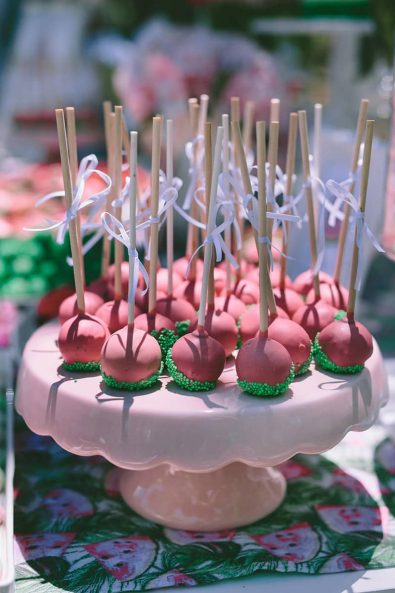 Watermelon inspired cake pops with red chocolate and green sprinkles and white ribbon ties on the lollipop stick for a baptism ceremony.
