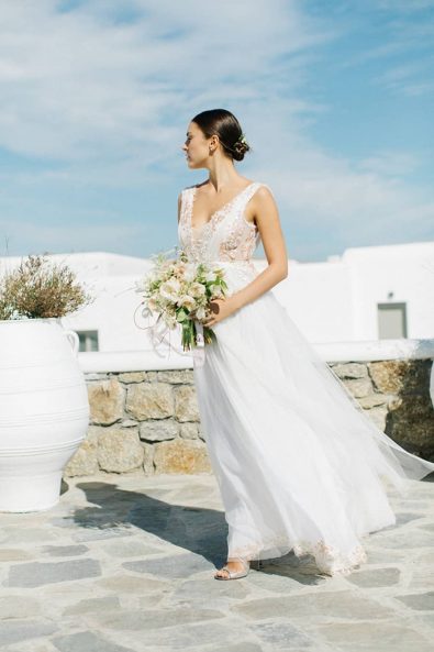 Chic and elegant bride wearing a romantic summer dress and blush bridal flowers on a mykonian house.