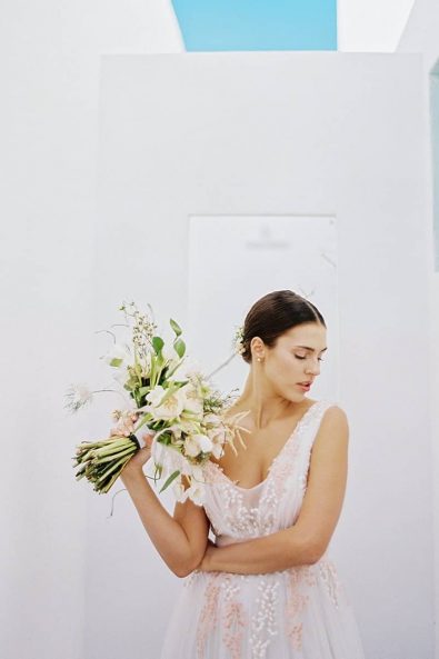 Minimal fine art bridal portrait, bride with a romantic wedding dress holding summer flowers in front of a white wall.