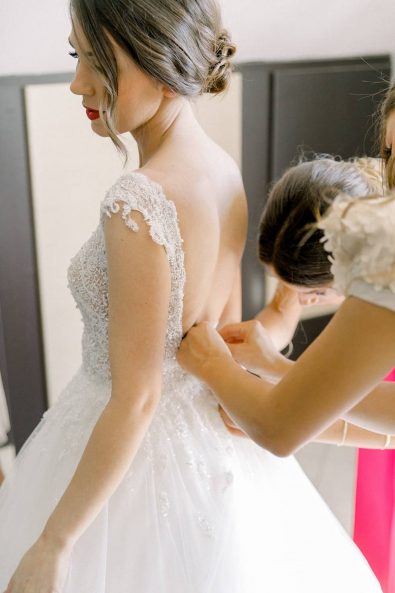 Bridesmaids help the bride put on her white lace, crystals and tulle wedding dress and tie the buttons in the back.