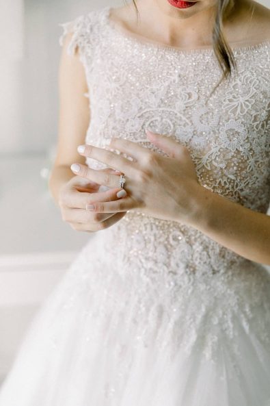 Detail shot of bride's hand slipping on her diamond wedding and engagement rings while she wears her romantic tulle gown and red lipstick.