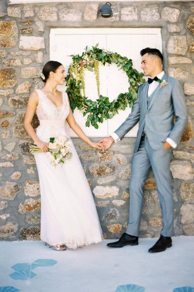 Lovely couple in front of a stone wall and a hanging wreath wedding flower decoration holding hands and looking at each other.