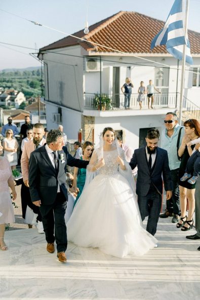 Bride walking to the church with the bridal parade, her father and her brother in a destination wedding in Messolonghi Greece.