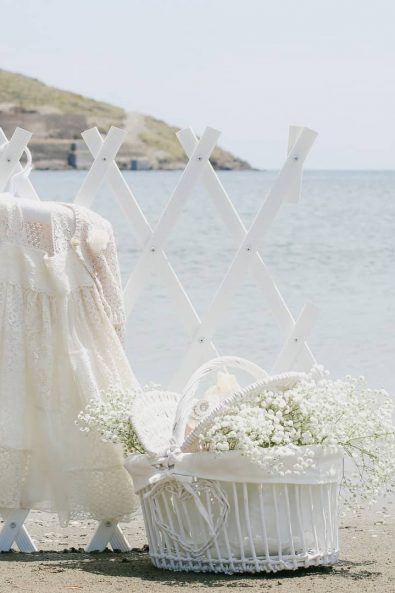 First communion christening white dress photographed on the beach next to a white basket full of baby's breath flowers.