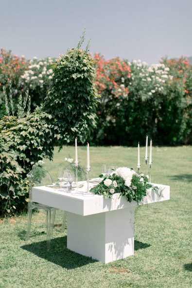 Bridal table on the garden with transparent chairs, vine background, and flower centerpiece.