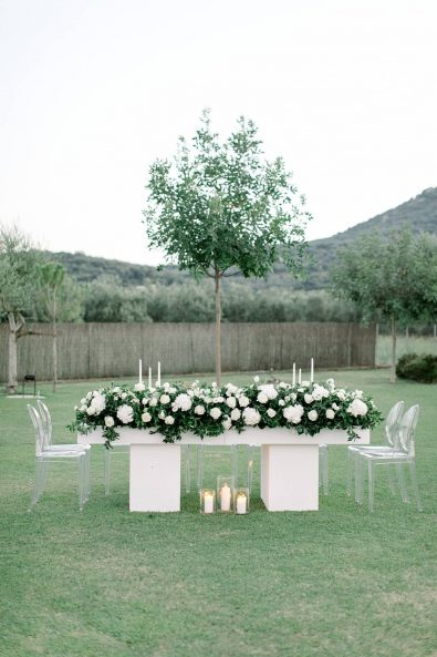 Wedding family table with white flower decorations, clear chairs, and candles on the grass.