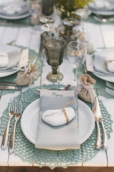 Teal crochet placemat, white porcelain plate, silver cutlery, dusty blue linen napkin, white stone wedding favor with silver fish, a burlap bag with ribbon and almond sweets, olive water glass and crystal wine glass.