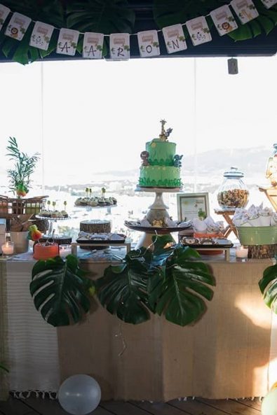 Baptism Party candy bar with birthday cake, cupcakes, biscuits, and marshmallows, decorated with green banana leaves, white and orange balloons.
