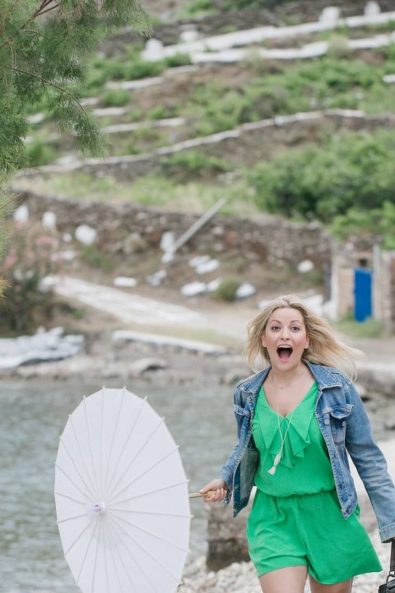 A blonde woman wearing a green playsuit and denim jacket and a man wearing a pink shirt and blue trousers laughing while walking on the beach.