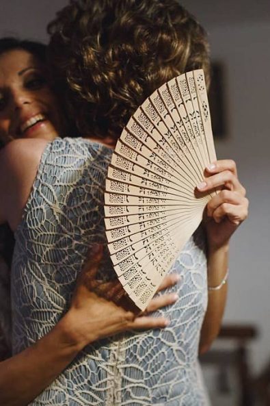 Happy bride holding a hand fan and hugging tightly her mother that wears a light blue lace dress.