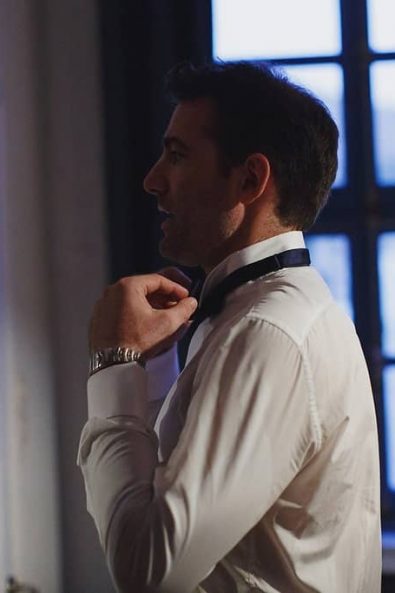 Groom in a white shirt getting ready tying his navy blue neck bowtie in front of a big blue window.
