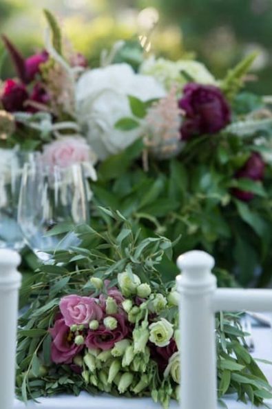 Close up of the bridal table with the white wooden chairs and the crystal glasses in the background and table flower arrangements with roses and peonies for a Greek-Arabic wedding in Greece.