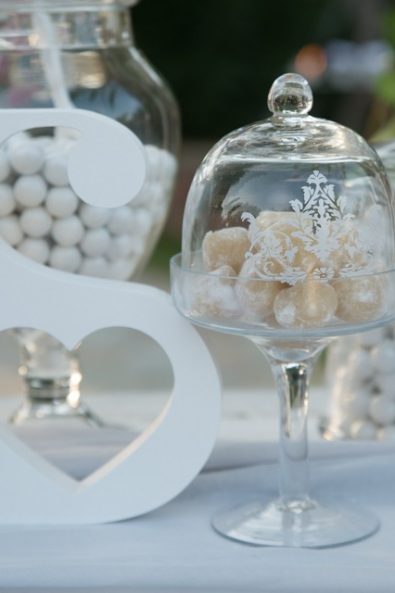 Glass pedestal cupcake stand with a dome, filled with almond wedding treats and white wooden initial decorative object.