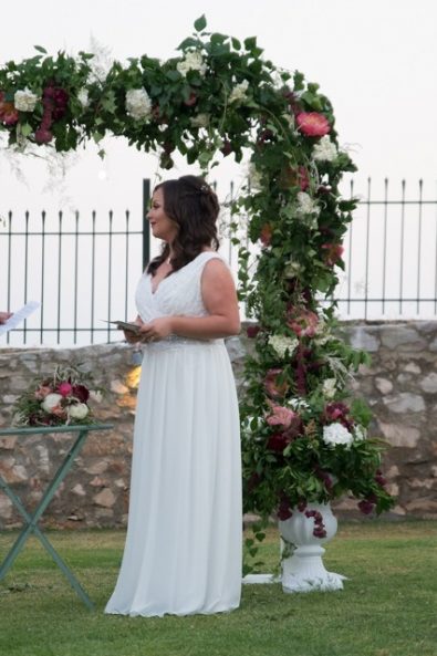 Beautiful bride with ancient greek hairstyle and white dress exchanging vows with her groom in front of the wedding arch.