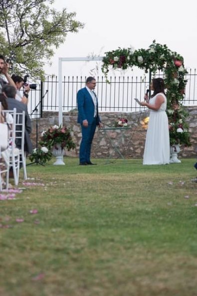 A couple exchanging vows in the afternoon under a white and deep red flowers decorated altar while the guests look at them.