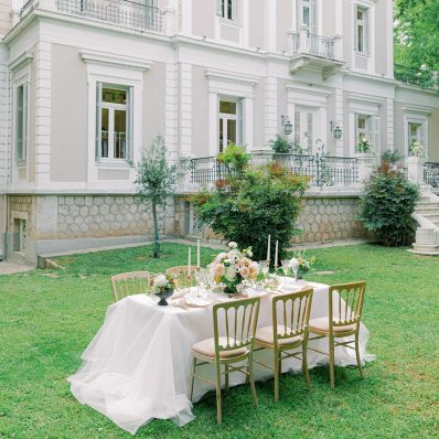Garden wedding area to elope in Greece, with a white clothed table with six chairs and culinary for dinner, in front of a white building.