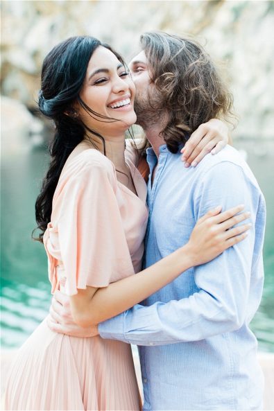 Groom kissing bride on cheek in front of Lake Vouliagmeni