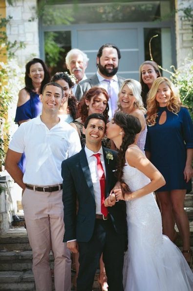 Family wedding portrait with family members, friends, groom and bride kissing his cheek.