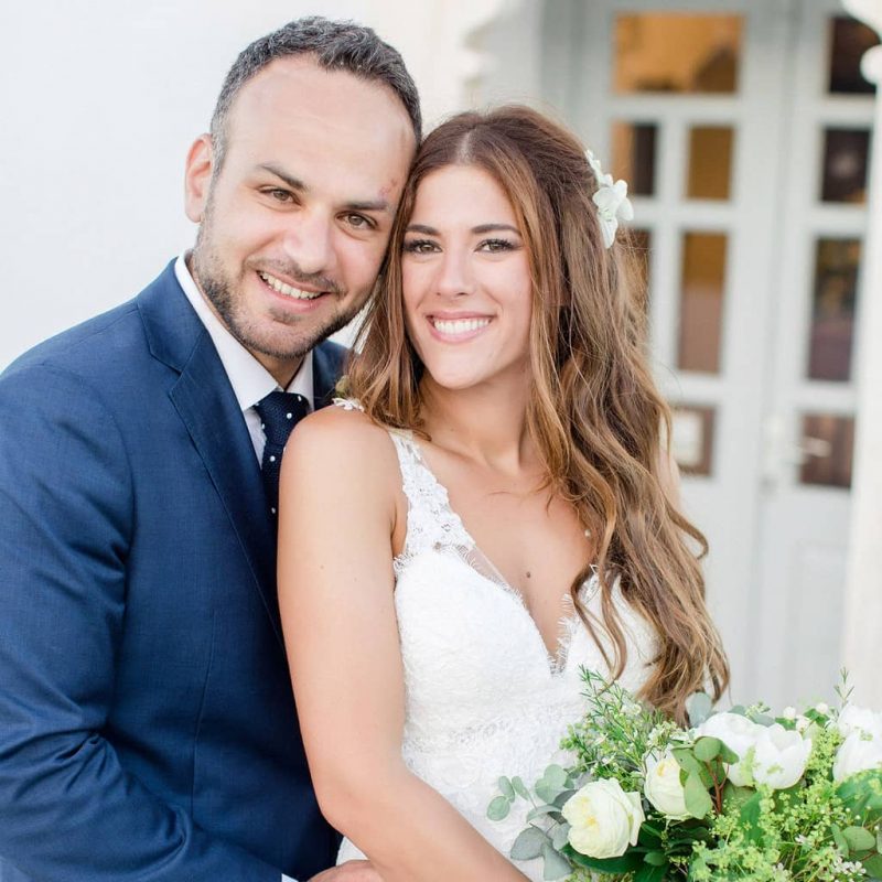 Beautiful young couple looking at the camera, groom with blue suit and polka dot neck tie and bride with mermaid hairdo and white flowers on the day of the wedding