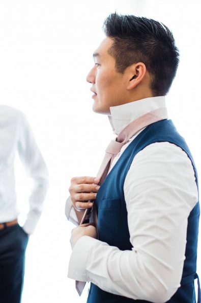 The groom in his navy blue suit getting ready with his friends tying his blush necktie.