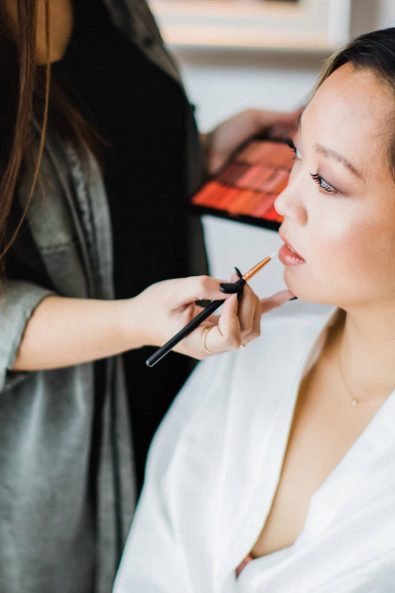 Bride in a silk white robe getting a minimal and romantic rosy bridal makeup by the makeup artist.