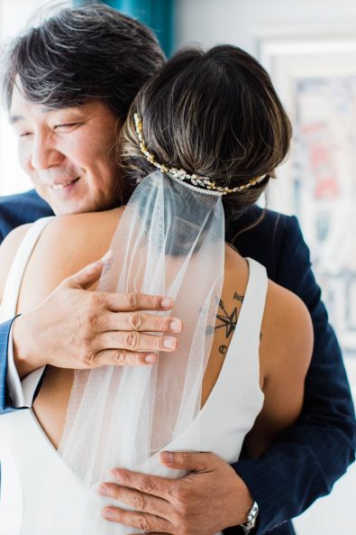 Bride in a minimal white dress and veil hugging her father before her destination wedding in Greece.