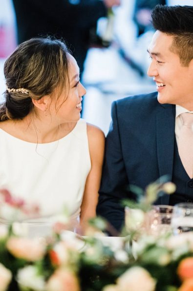 The bride and the groom sitting at the wedding table and looking lovingly into each other's eyes at a Korean destination wedding in Greece.