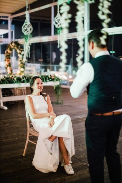 Groom at a wedding party reception ready to remove the bride's garter custom while she sits in a wooden chair at their destination wedding in Greece.