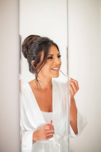 The bride putting on her mauve bridal lipstick in front of a large white mirror wearing a silk robe and nightgown.