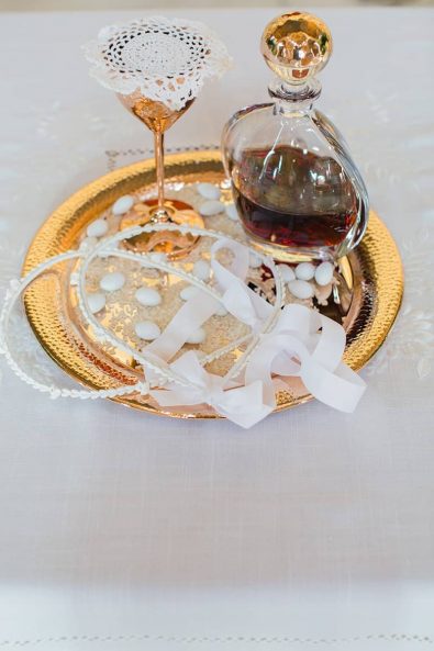 Close up shot of gold tray with sugar-coated almonds and rice, vintage white wedding crowns, red wine glass and copper wine goblet for the Orthodox wedding ceremony.