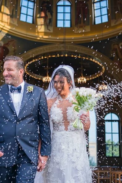 The bride and groom holding hands exiting the church of their wedding while the guests are tossing rice to them for good fortune and fertility after their destination wedding in athenian riviera.