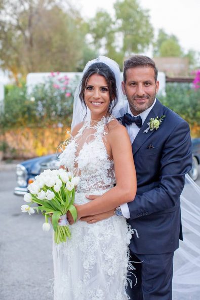 Photo of a bride in a sexy wedding dress and groom navy blue suit posing in a warm embrace while they look at the camera after their destination wedding in athenian riviera .