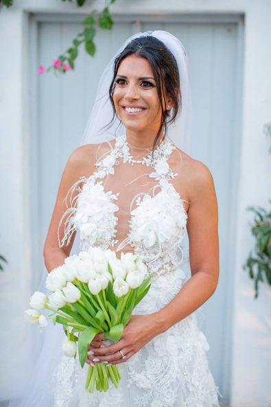 Just married bride looking very happy in her lace and feathers sexy wedding dress and silk tulle veil, holding her white tulips bridal bouquet at a destination wedding in athenian riviera.