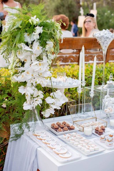 Close up shot of dessert station candy bar dessert stand in white and grey, with white orchid flower arrangements, marbled meringues, and copper brushed macarons, chocolate cupcakes, custom made sugar biscuits and white candles.
