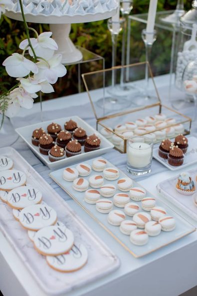 Close up shot of candy bar with custom made wedding biscuits with couple's initials, white copper brush decorated macarons, marbled cream puffs and meringues, and mini chocolate cupcakes.