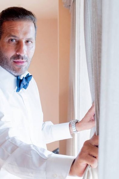 Groom wearing a crisp white shirt and blue bow tie in front of a window.