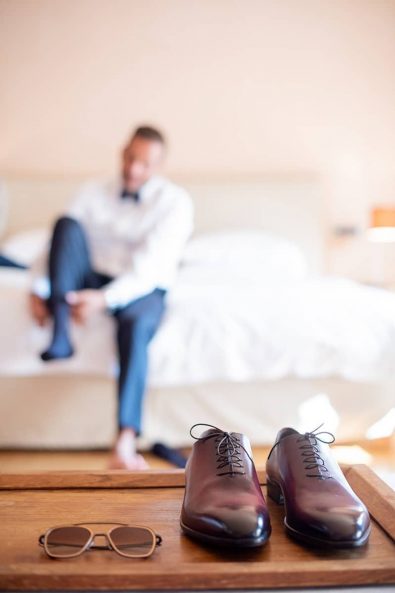 Close up shot of groom's shoes and glasses with him in the background getting ready.